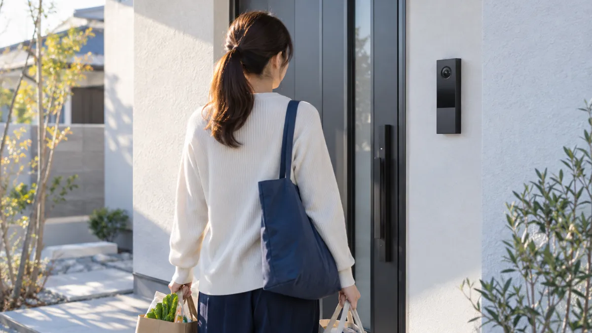 夕方の戸建て住宅の玄関前で、買い物袋を持った人が玄関横の縦長の顔認証インターホンを見ている後ろ姿