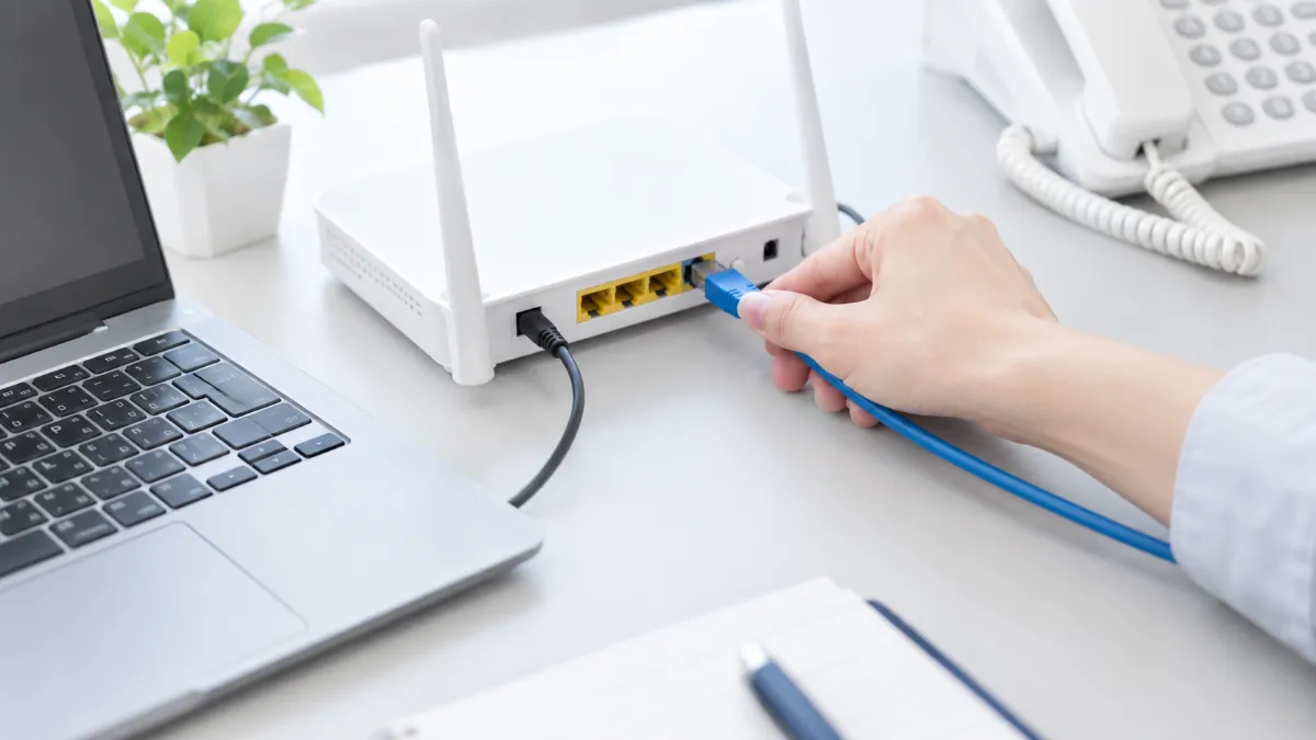机の上で、ノートパソコンの横の Wi-Fi ルーターに LAN ケーブルを差し込んでいる手元と、奥の固定電話機